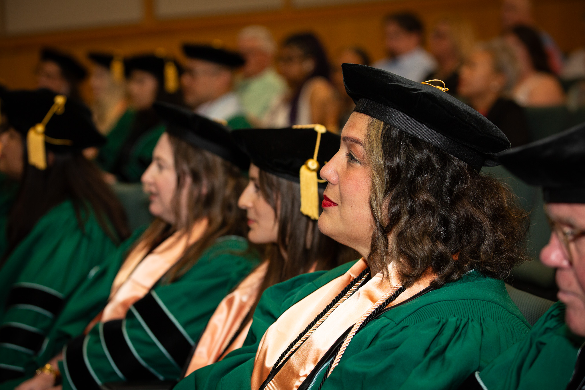 Three public health doctoral candidates dressed in commencement regalia, listening with a large audience