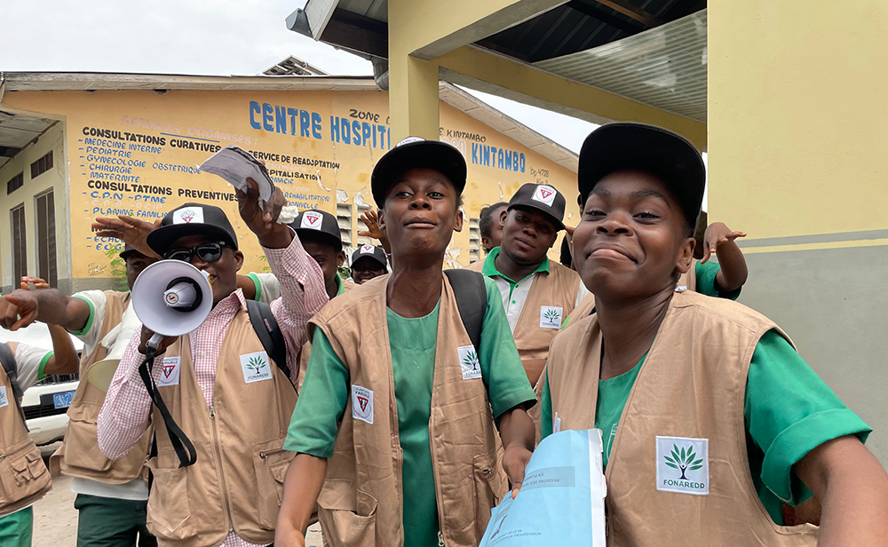 Group of young Black men smiling, wearing vests with organization badges, standing outside of a Health Centre in Kintambo