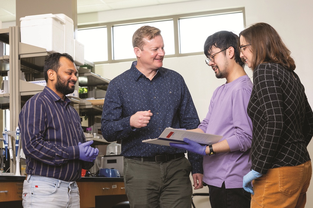 Four smiling scientists discuss work in a lab, one holding papers.