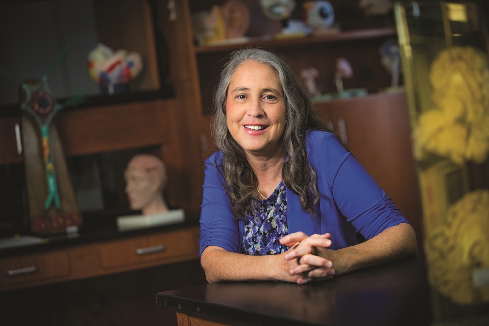Smiling woman at a desk in a science lab, surrounded by anatomical models.