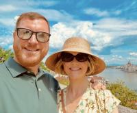 Chris Richardson and Andrea Schippert Richardson smiling with a European city and river in the sunny background.